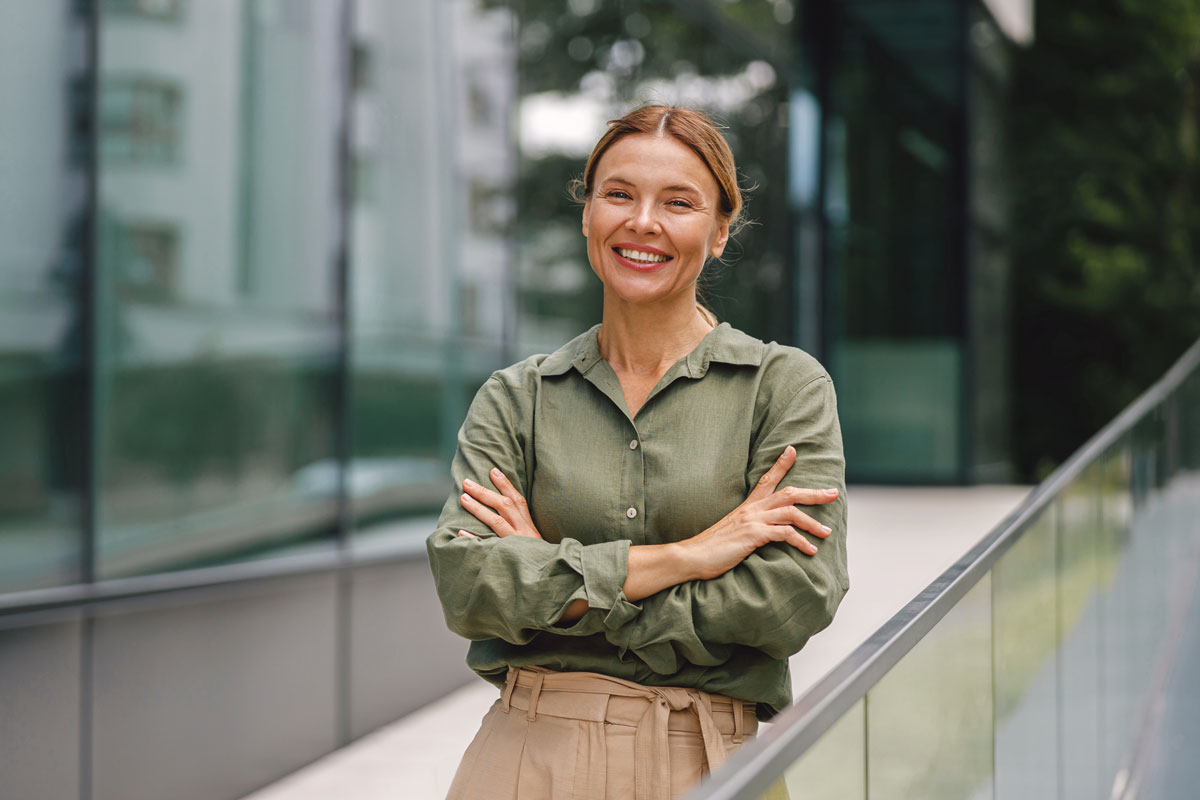 Woman with rolled-up sleeves and crossed arms leaning on a railing in front of a modern glass building