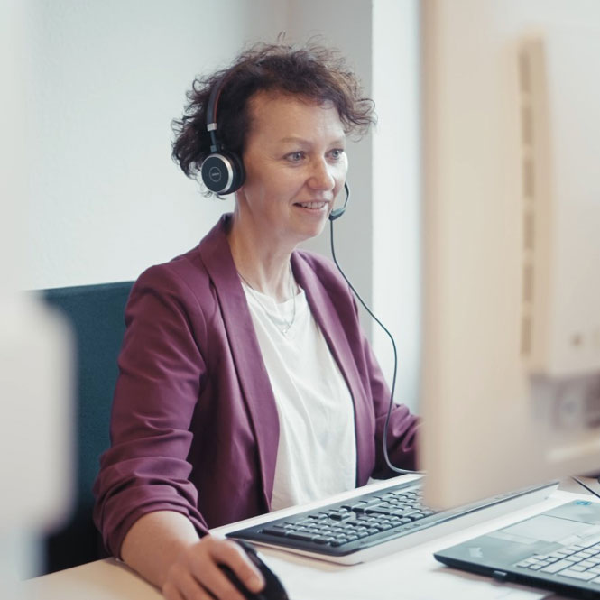 Frau mit Headset sitzt an einem Schreibtisch und arbeitet am Computer in einem Büro.