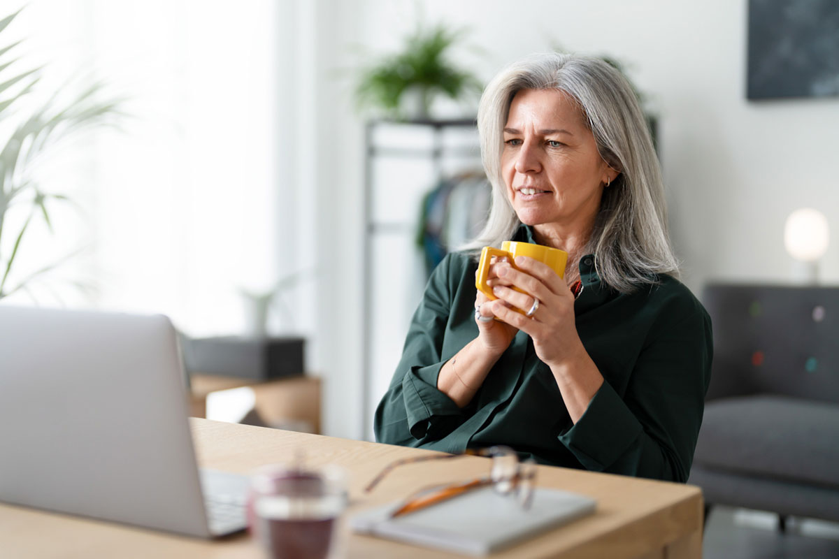 Older woman with gray hair sitting at a table holding a yellow mug with both hands