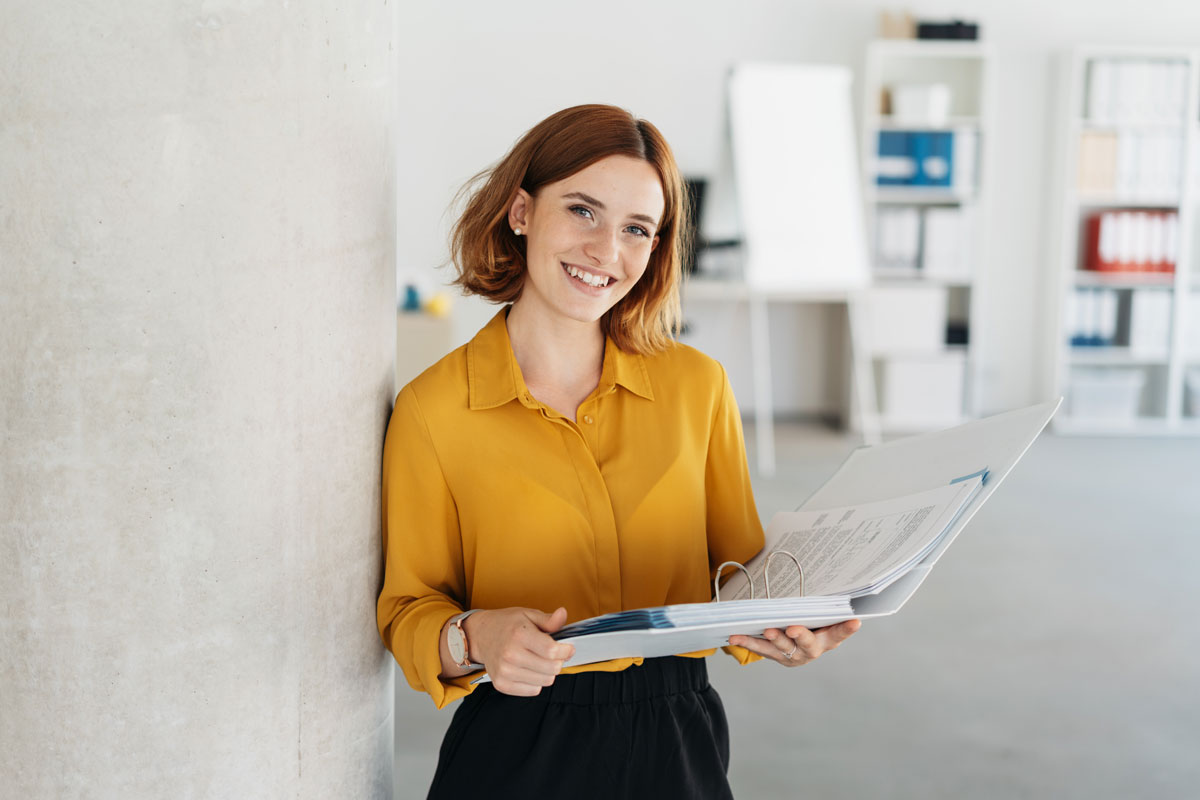 Woman with shoulder-length hair wearing a yellow shirt leaning against a wall holding an open binder in a bright office.