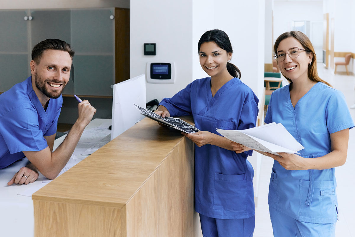 Three people in medical scrubs stand at a reception desk, two holding papers, one leaning on the counter with a pen at the mouth.